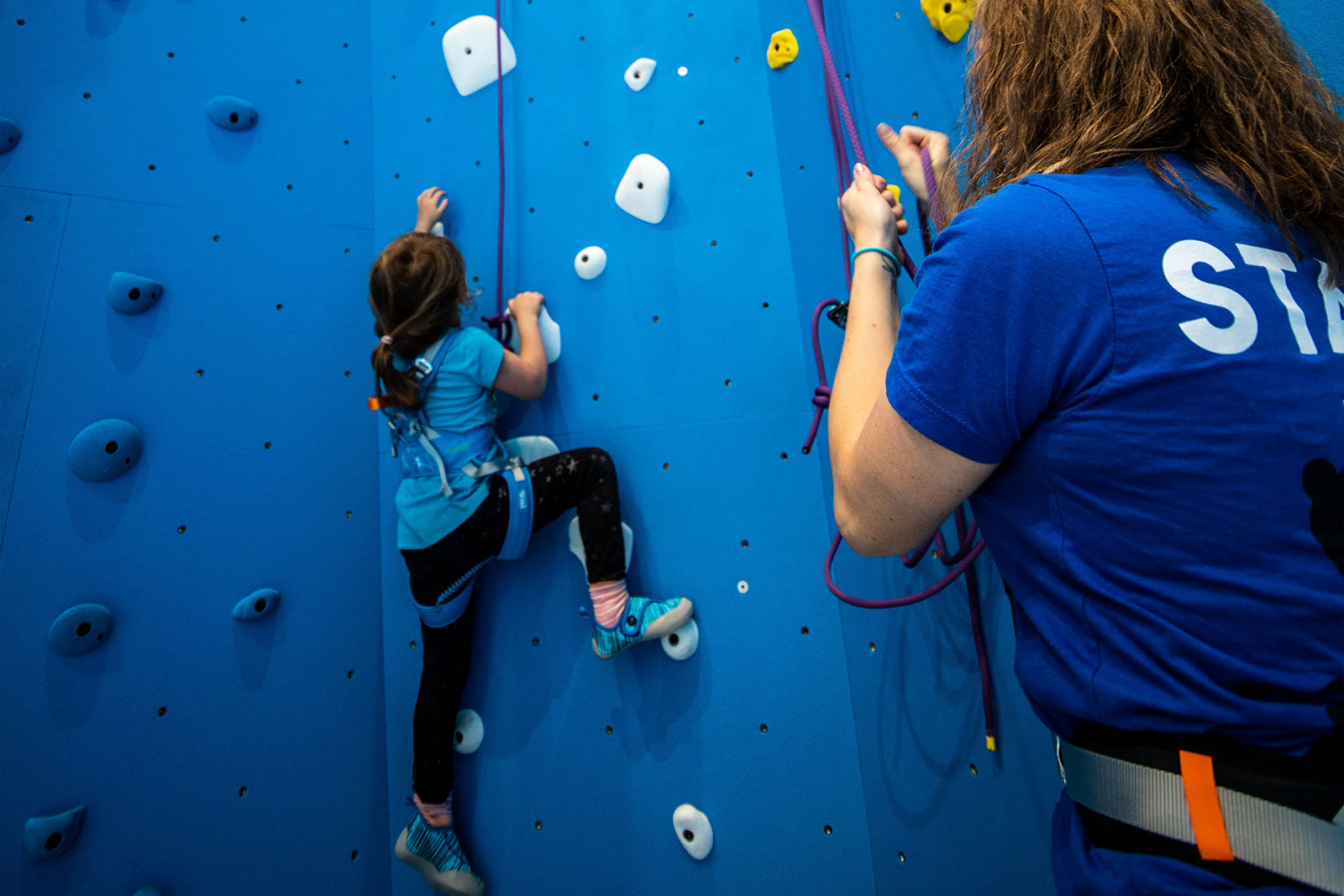 child climbing on blue wall being belayed by an instructor