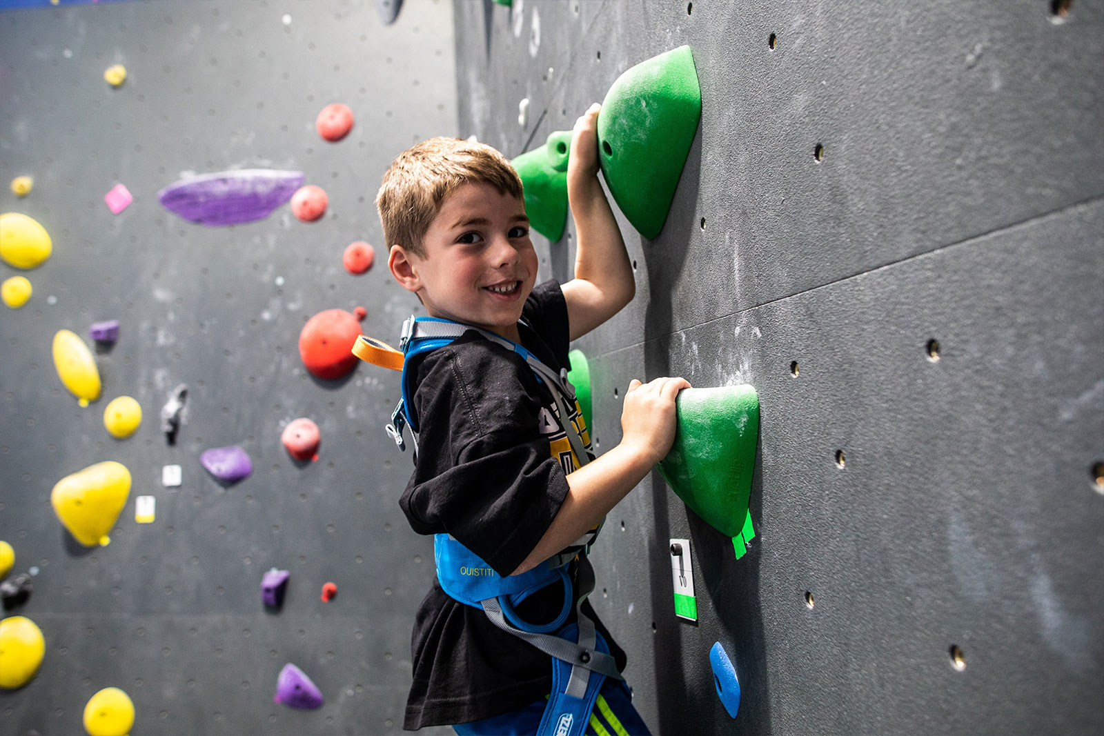 child holding onto rock green grips on gray rock climbing wall smiling at camera